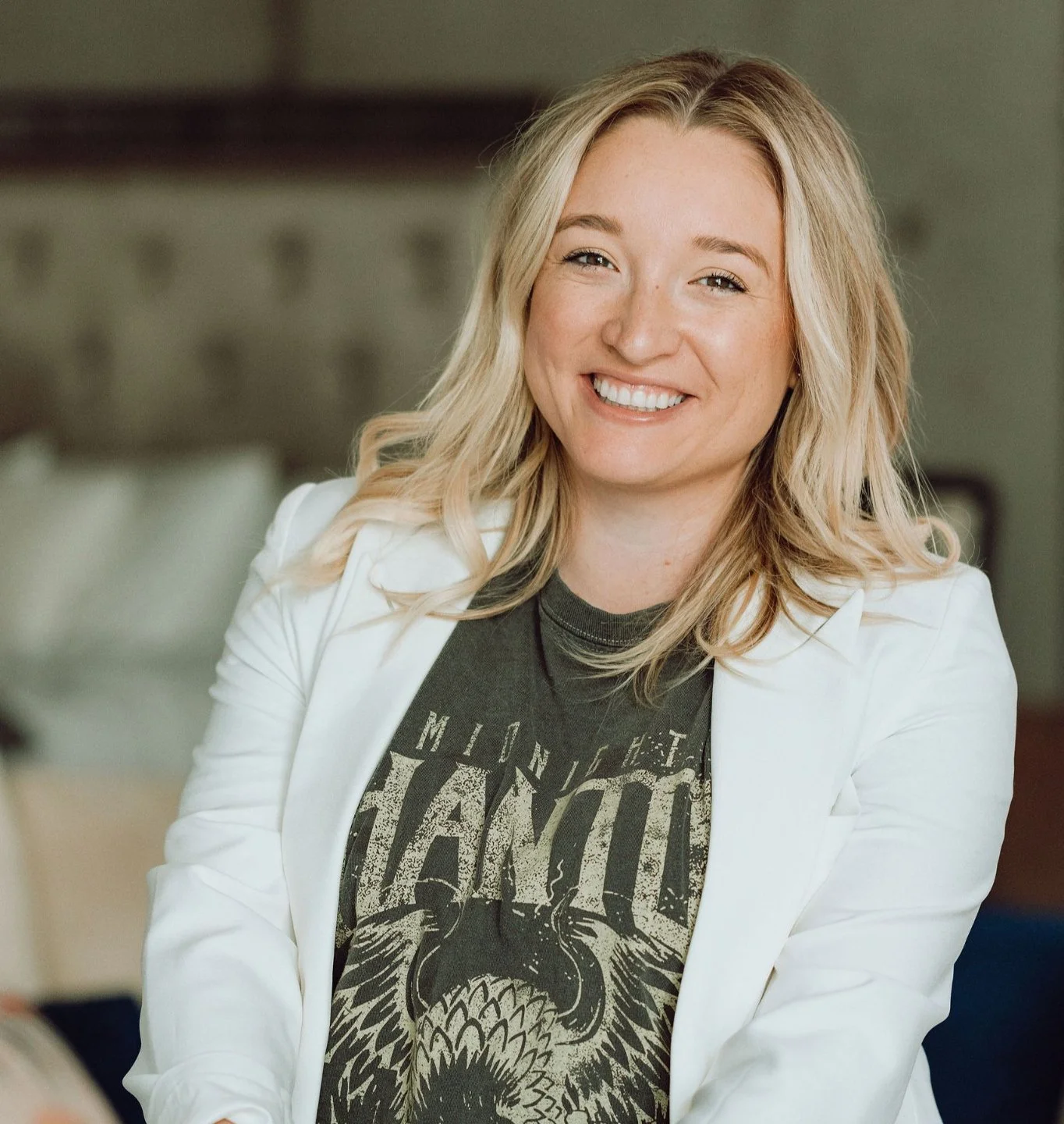 A person with long blonde hair, smiling and wearing a white blazer over a graphic T-shirt with the text "MIDNIGHT PHANTOM."