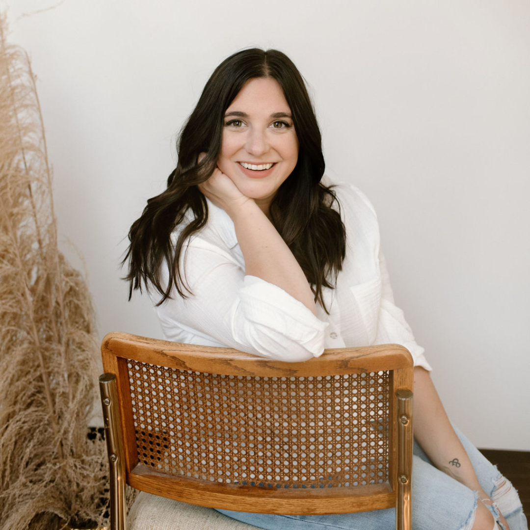 Woman with long dark hair, wearing a white shirt, sitting on a wooden chair, smiling, with dried plants in the background.