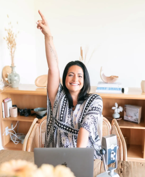 Smiling woman sitting at a desk, raising her arm, with a laptop and microphone in front.