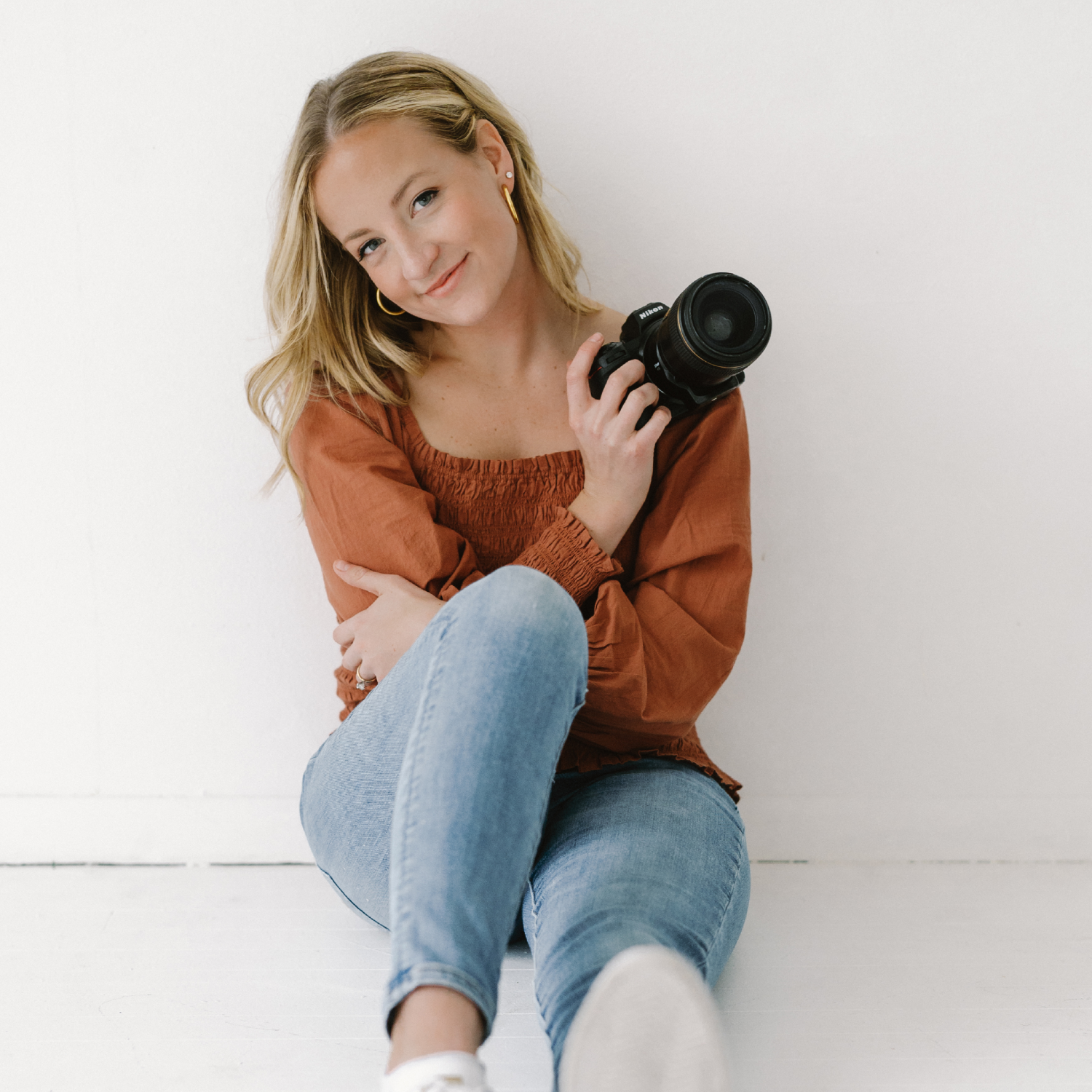 Person sitting on floor holding a camera, wearing a rust-colored top and jeans against a white background.