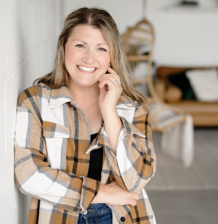 Person smiling, wearing a plaid shirt, standing against a white wall in a bright room with a chair and couch in the background.