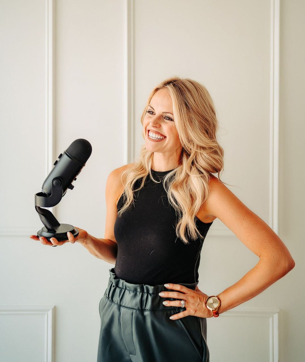 Woman holding a microphone, smiling, wearing a black sleeveless top and green pants, standing indoors.