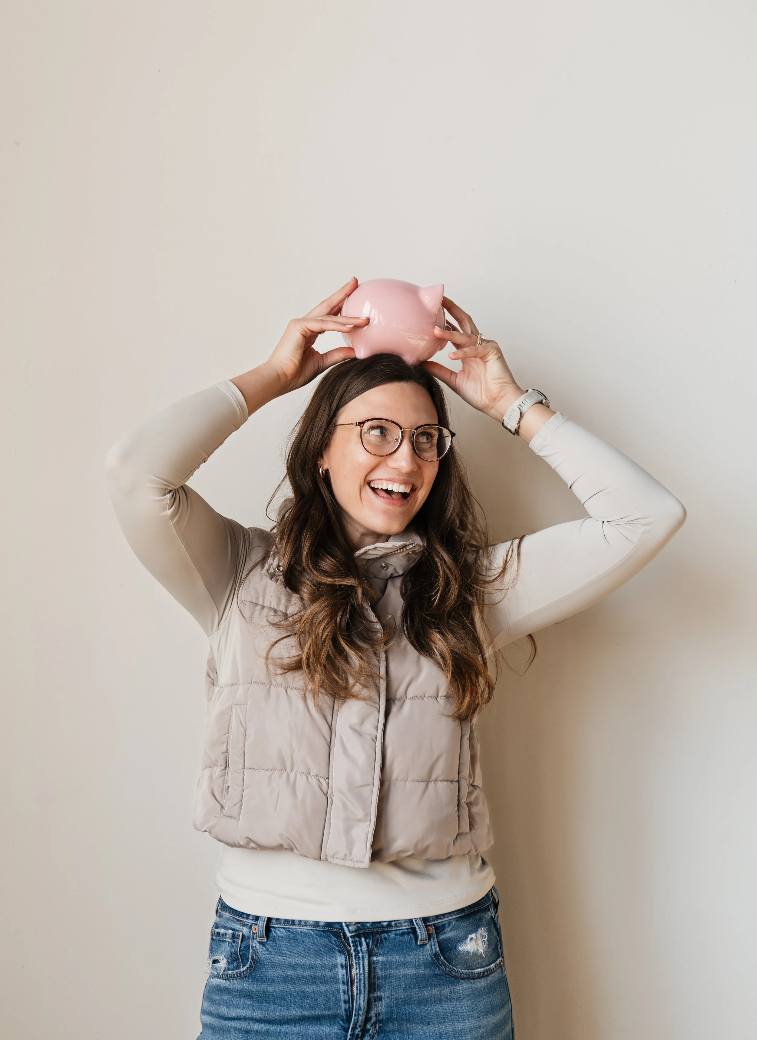 Woman smiling, holding a pink piggy bank above her head, wearing glasses, cream long-sleeve shirt, and a beige puffer vest.