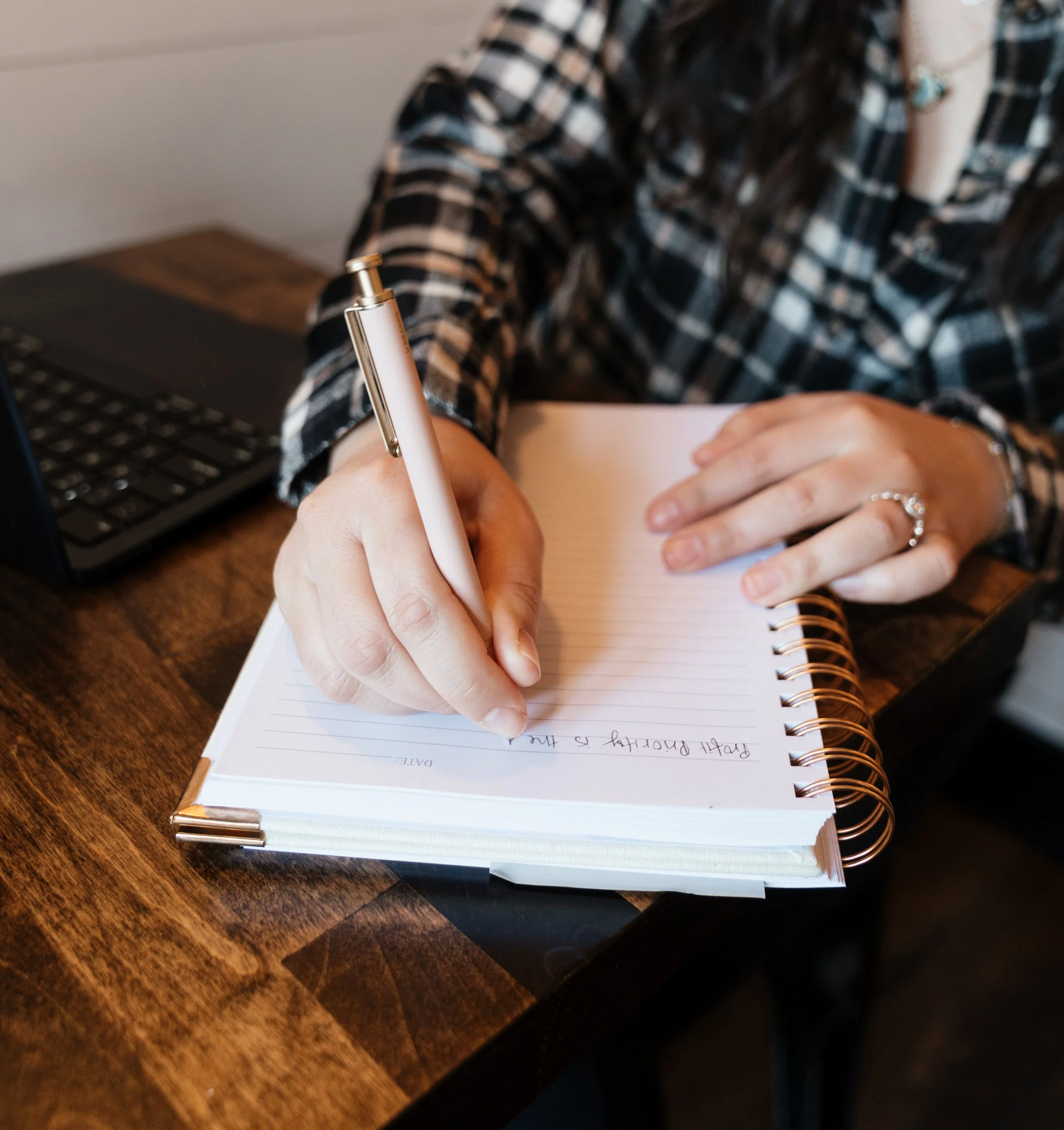 Person writing in a spiral notebook on a wooden desk with a laptop nearby.