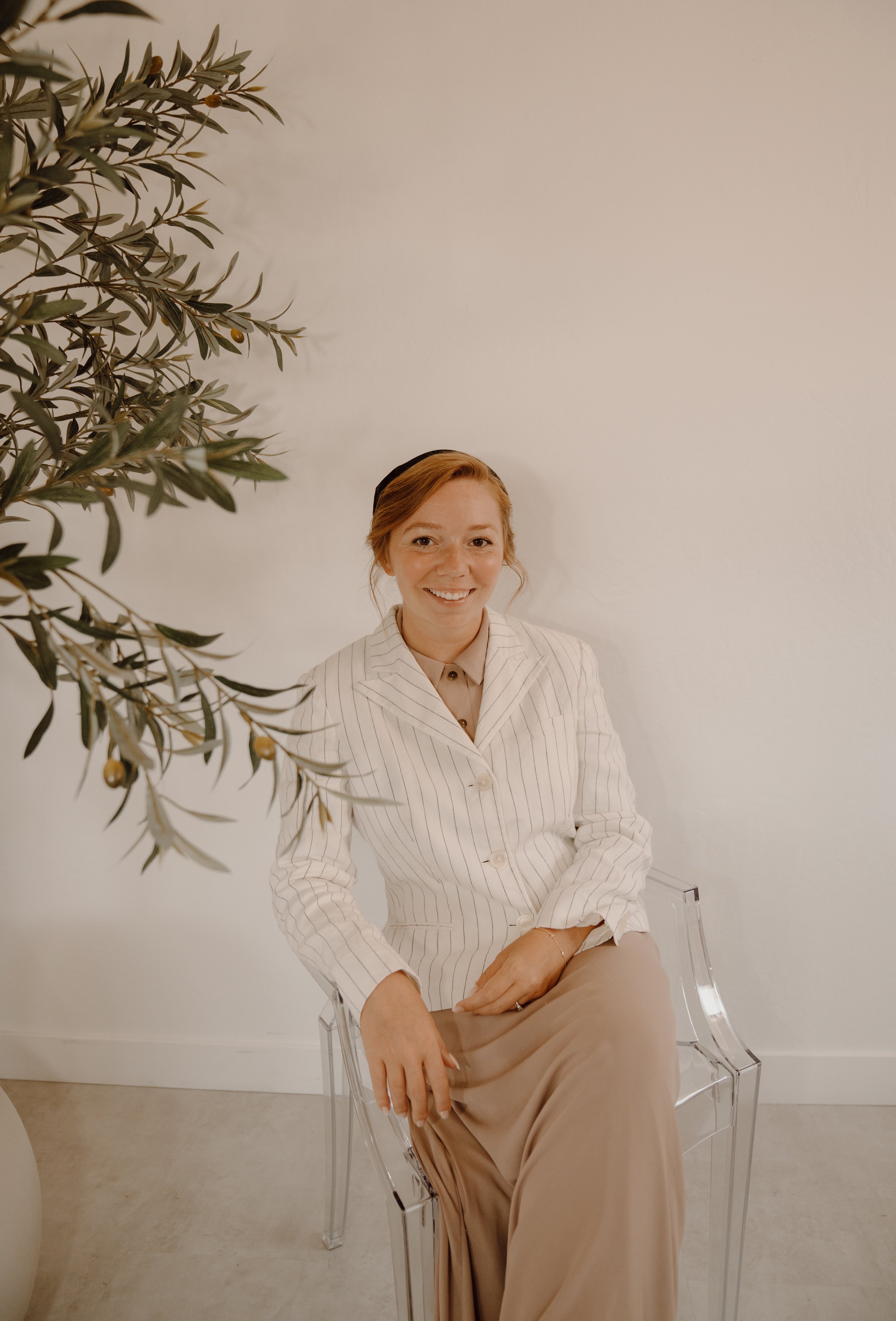 A person wearing a striped jacket and long skirt sits on a clear chair near an olive tree, smiling.
