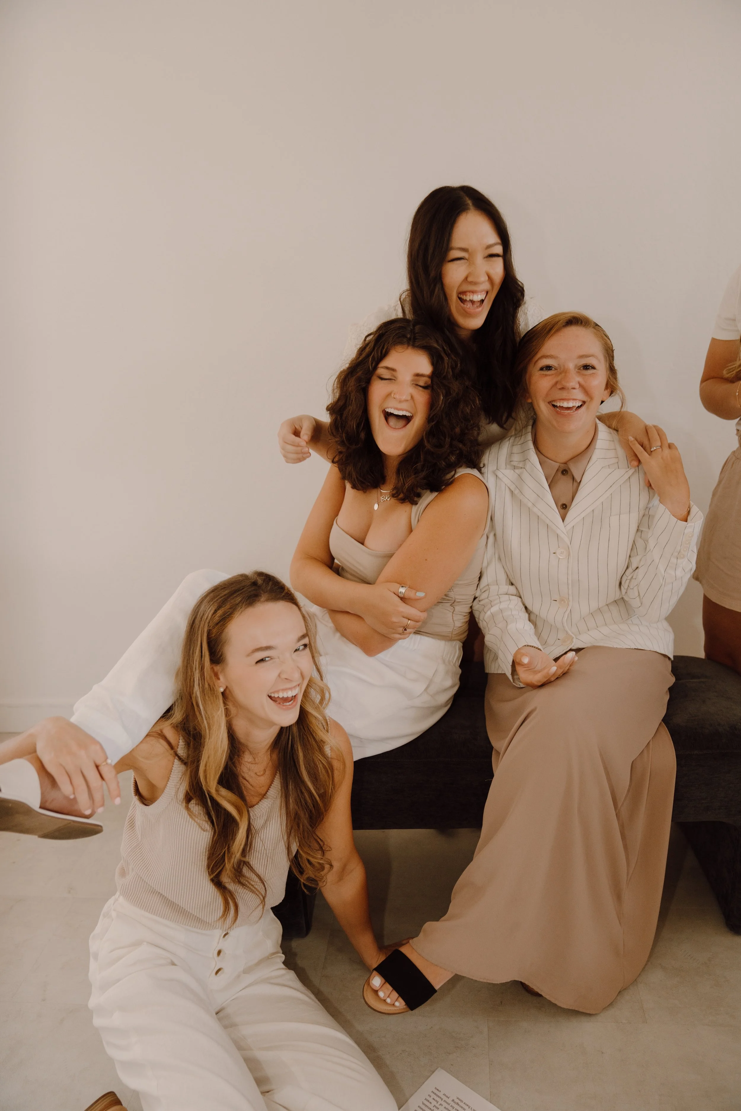 Group of four women laughing and enjoying each other's company, sitting and kneeling in a relaxed setting; they are wearing casual, neutral-colored outfits.