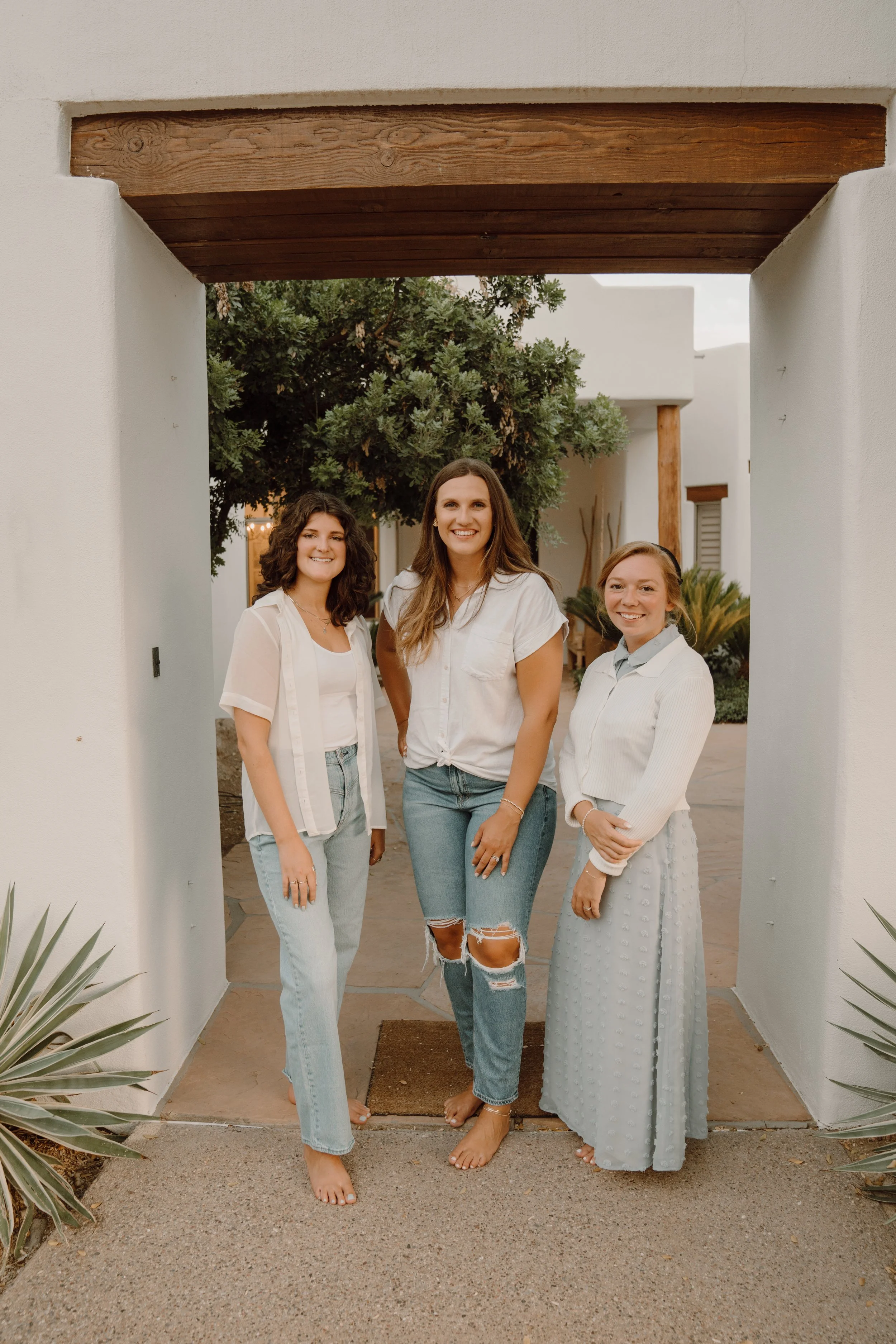 Three women standing barefoot under a white archway, wearing casual white tops and jeans or a long skirt. They are outdoors with a tree and modern building in the background.