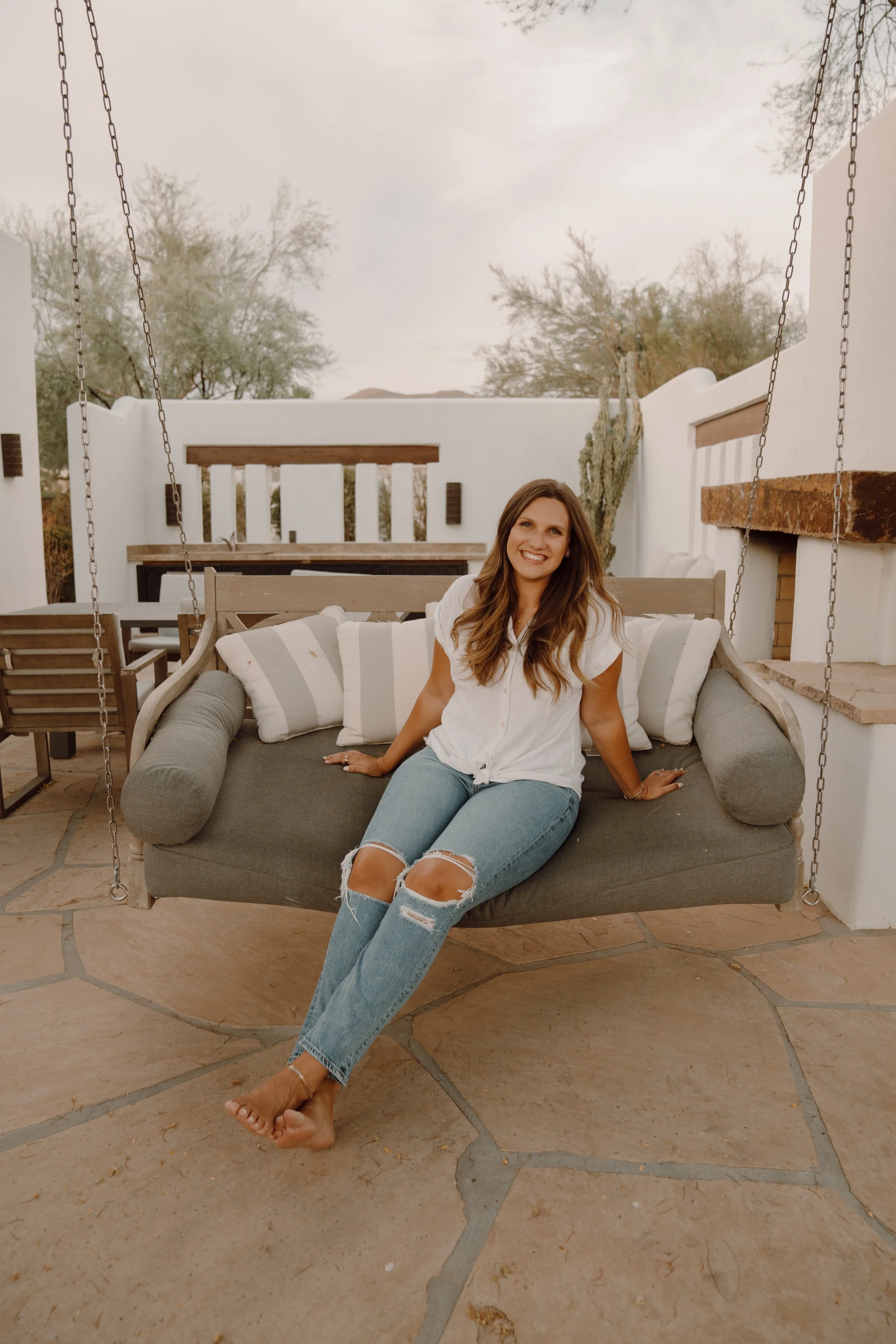 A woman sitting on an outdoor swing chair in a patio area, wearing a white shirt and ripped jeans, surrounded by a comfortable, stylish setting with a table, chairs, and greenery.