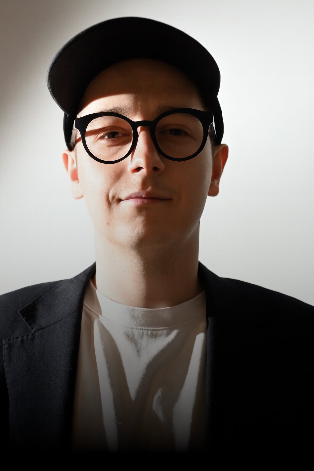 A young man wearing a black cap, black glasses, a black blazer, and a white T-shirt, looking at the camera against a plain light-colored background.