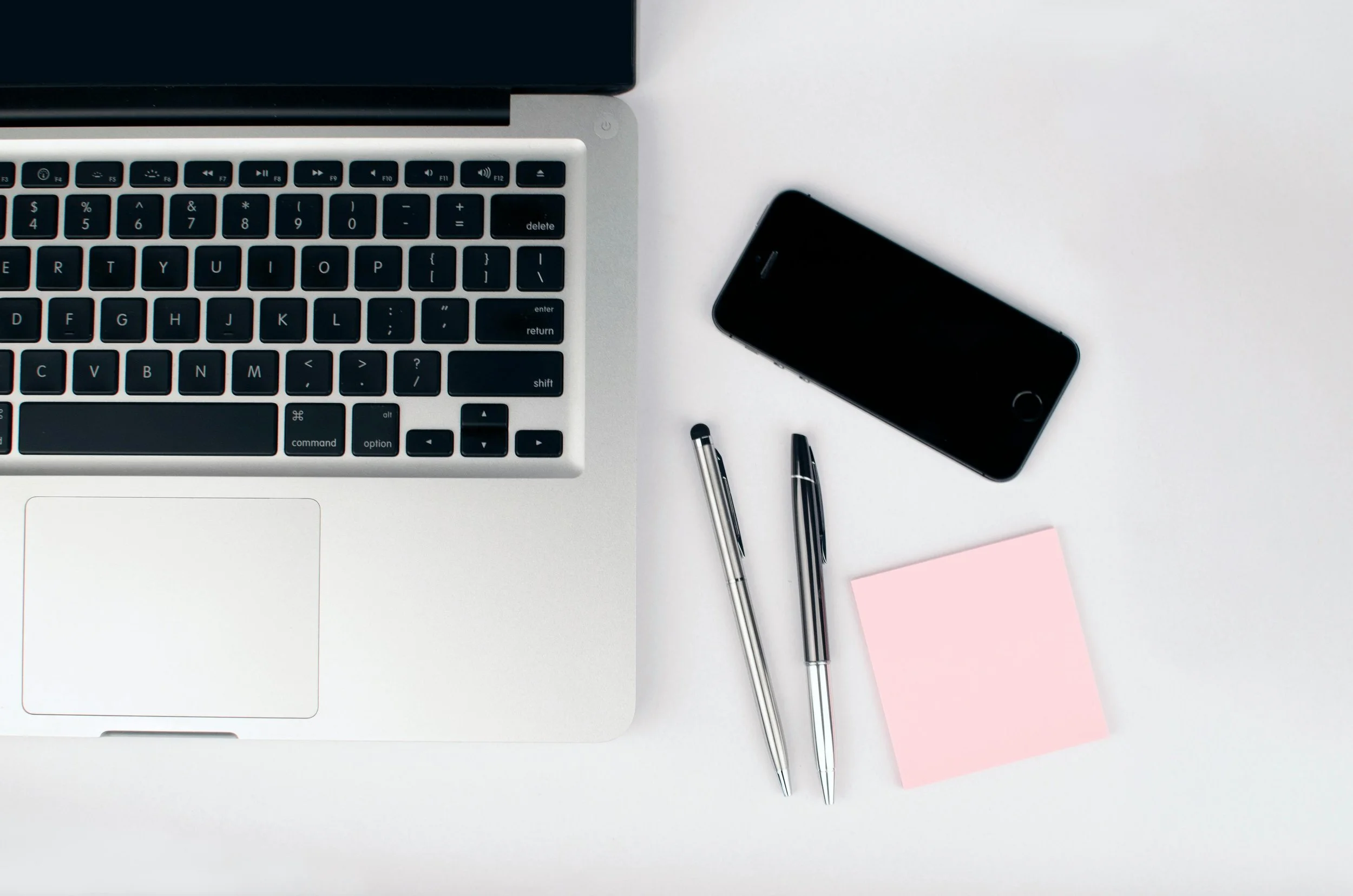 Top view of a workspace with a silver laptop, black smartphone, two silver pens, and a pink sticky note on a white surface.