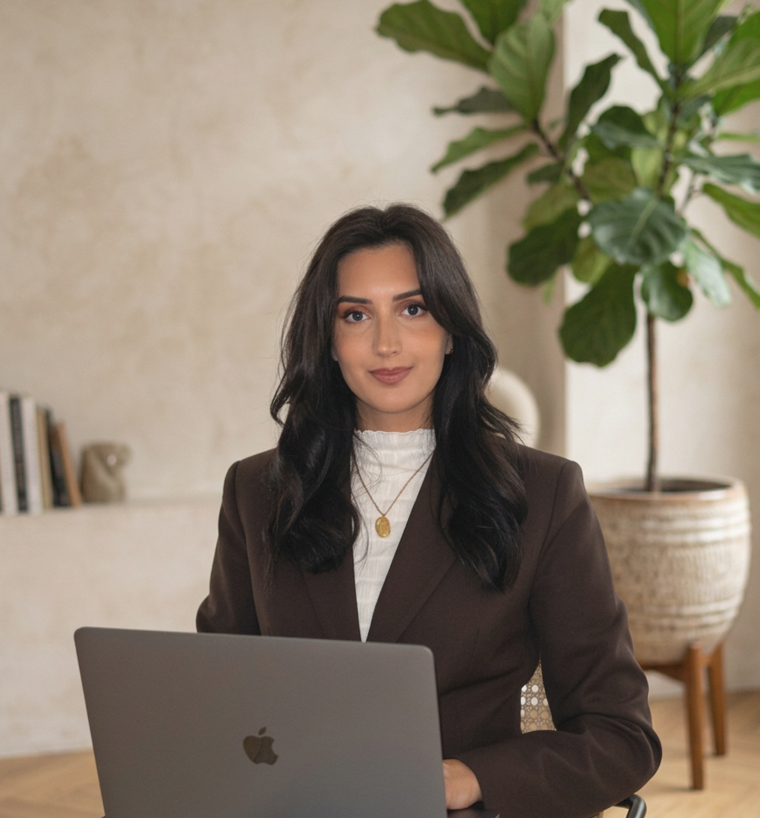 Salomé Dupin : femme assise à un bureau avec un ordinateur portable, dans une pièce décorée avec une grande plante verte et des livres en arrière-plan.