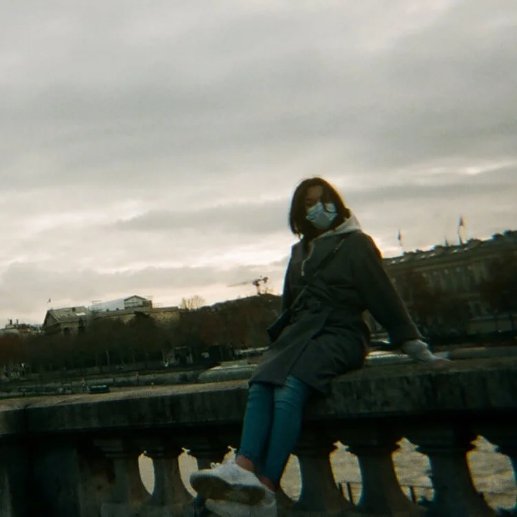 Une femme portant un masque facial, une veste sombre, des jeans bleus et des baskets blanches, assise sur une balustrade en pierre au bord de l'eau, avec un ciel nuageux en arrière-plan.