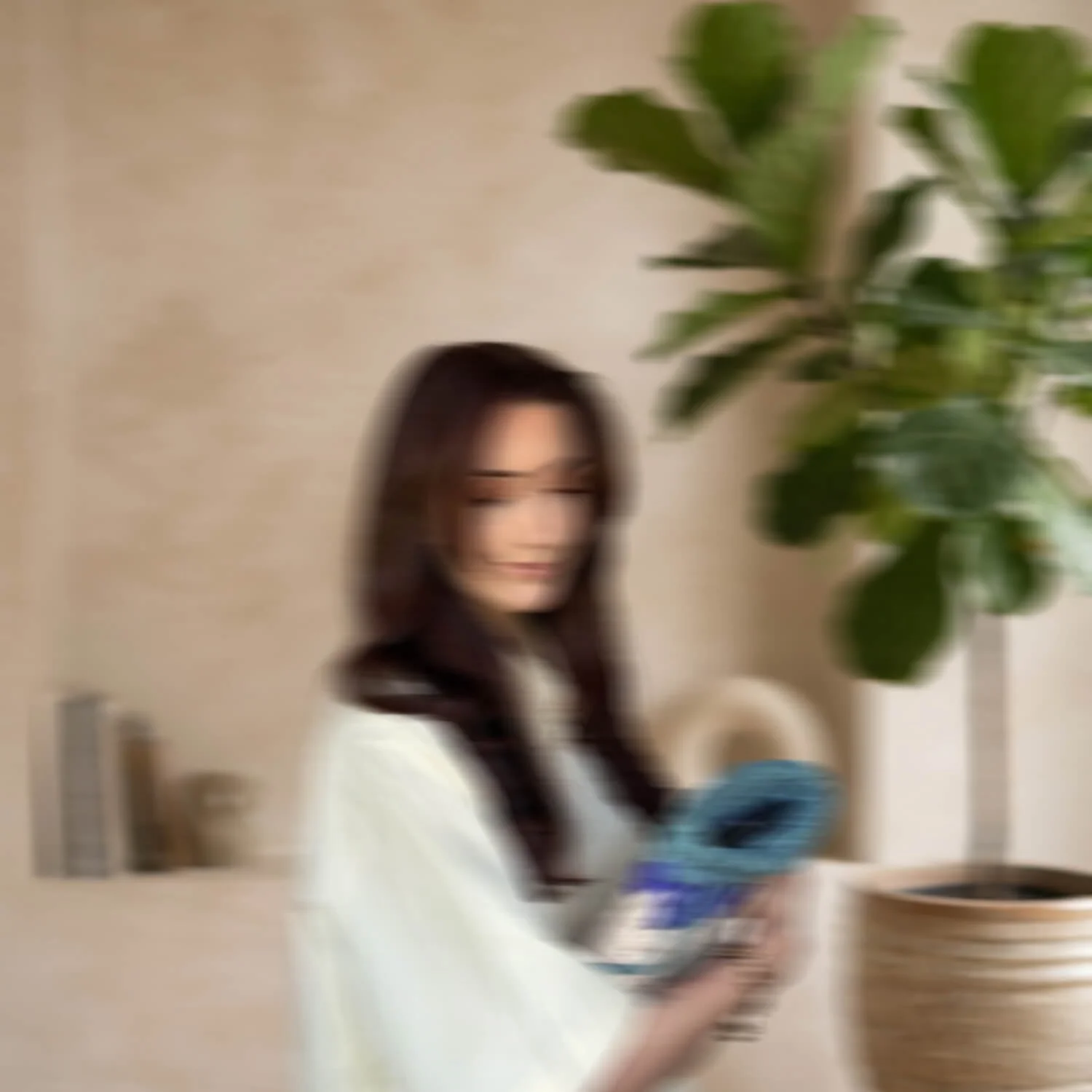Une jeune femme avec des cheveux longs et foncés tient une bouteille d'eau. Elle est assise devant un arbre d'intérieur avec de grandes feuilles vertes, le tout dans une pièce lumineuse.