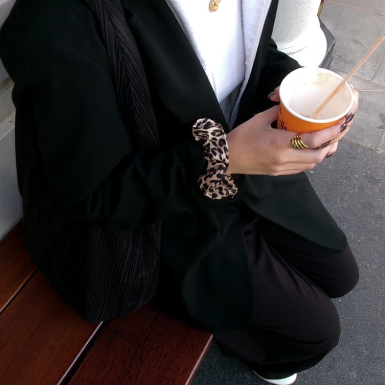 Une personne assise sur un banc en bois, tenant un pot de glace avec une cuillère, portant une veste noire et un pull gris, avec un accessoire en fausse fourrure à motif léopard au poignet, et plusieurs bagues.