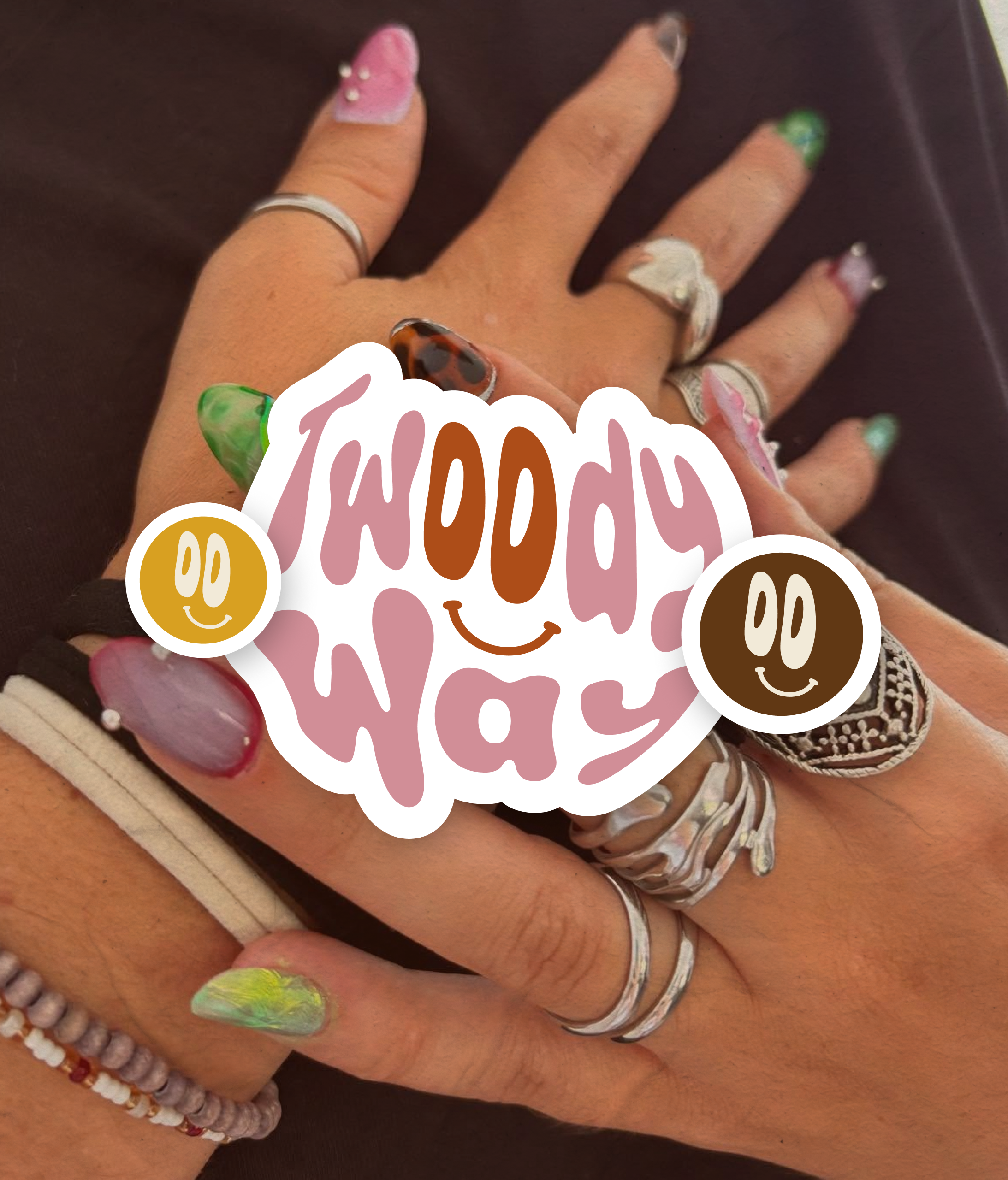 Une main portant plusieurs bagues et bracelets, avec des ongles peints de différentes couleurs et motifs, posée sur une surface sombre, avec un autocollant "Happy WDay" couvrant une partie de la main.