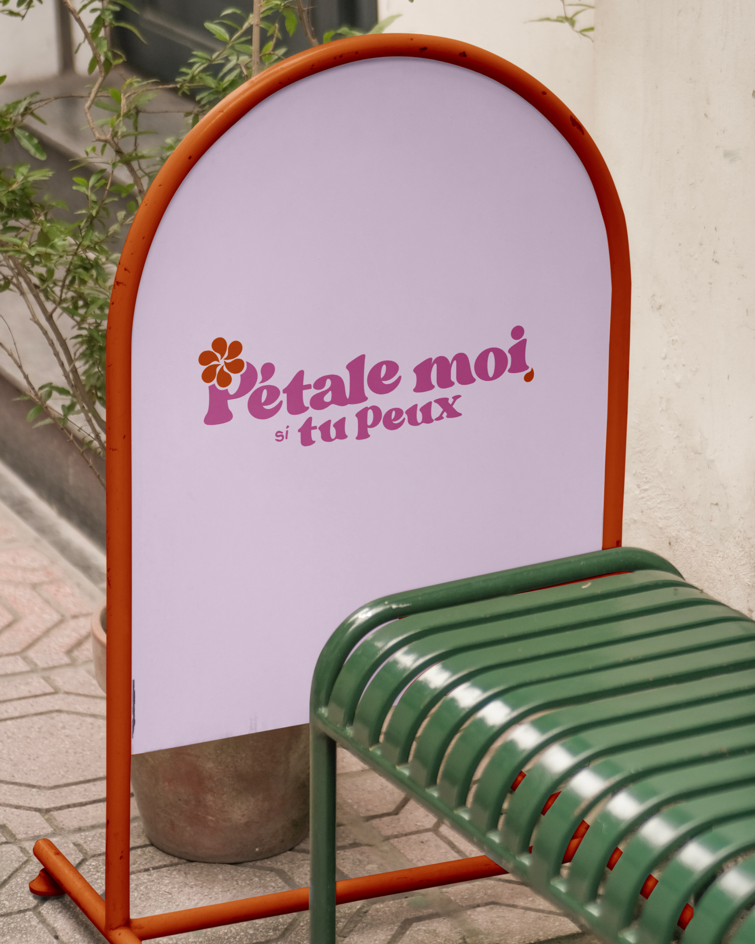 Panneau publicitaire coloré avec texte en français et une fleur, situé sur un trottoir et accompagnant un banc vert.