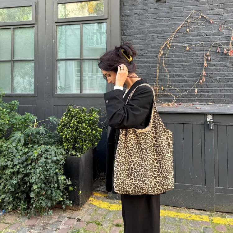 Salomé Dupin : femme avec une tenue noire, portant un sac en léopard, se tenant à côté de plantes en pot sur un trottoir en pierre, avec un mur noir en arrière-plan.