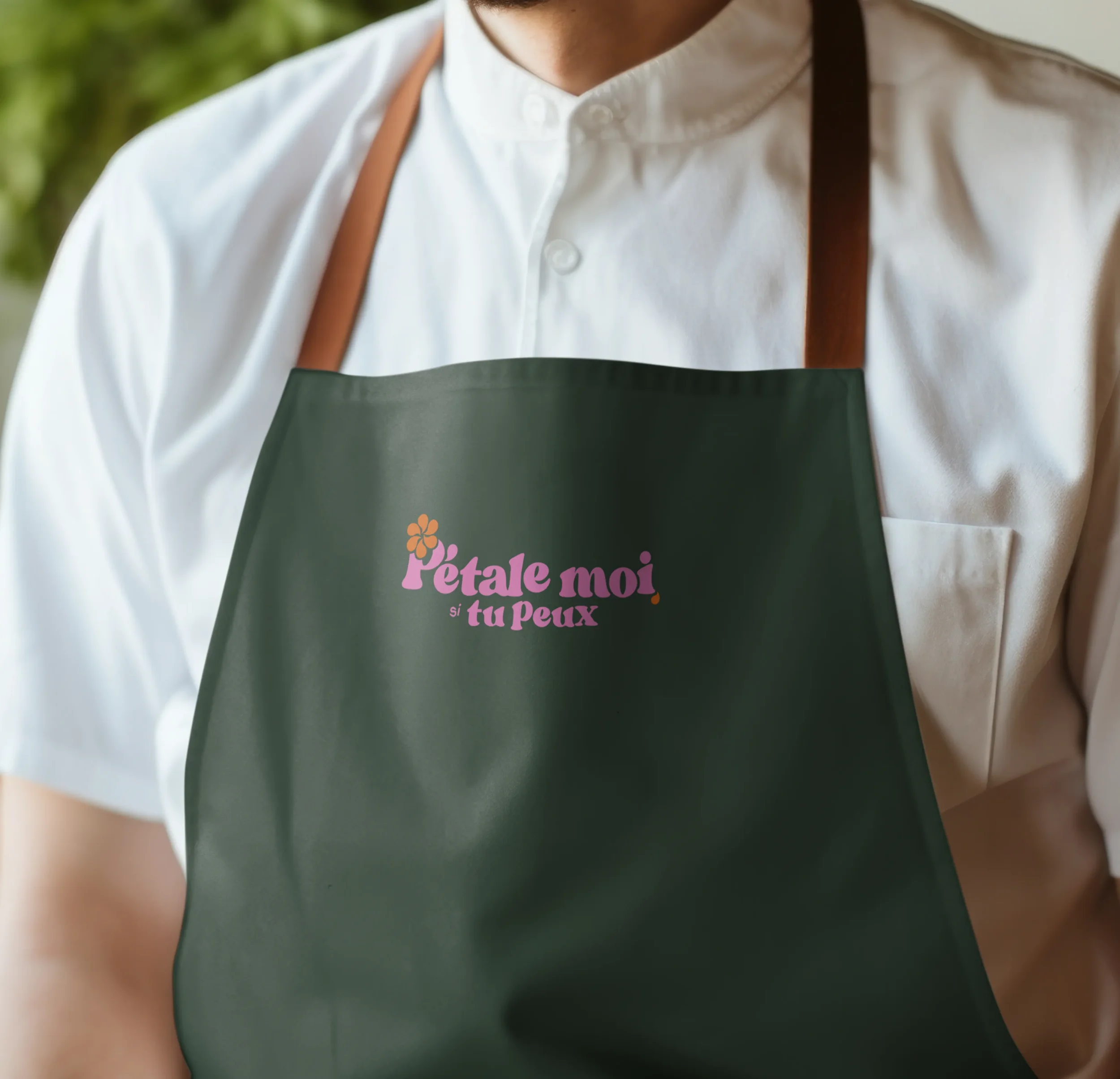 Un homme porte un tablier vert avec le logo 'Pétale moi si tu peux' en rose et orange, en train de cuisiner ou de servir dans une cuisine ou un cadre extérieur
