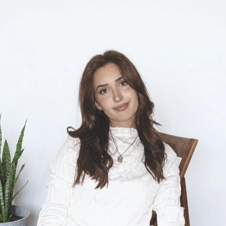 Salomé Dupin, une jeune femme aux cheveux bruns bouclés assise sur une chaise en bois, portant une blouse blanche, avec un collier, à côté d'une plante verte, sur fond blanc.