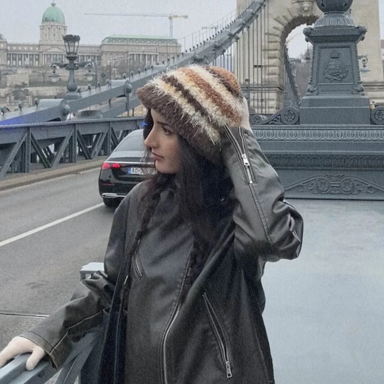Jeune femme avec un chapeau en fourrure et un manteau en cuir, en train de rouler un scooter sur un pont dans une ville, avec des bâtiments historiques en arrière-plan.