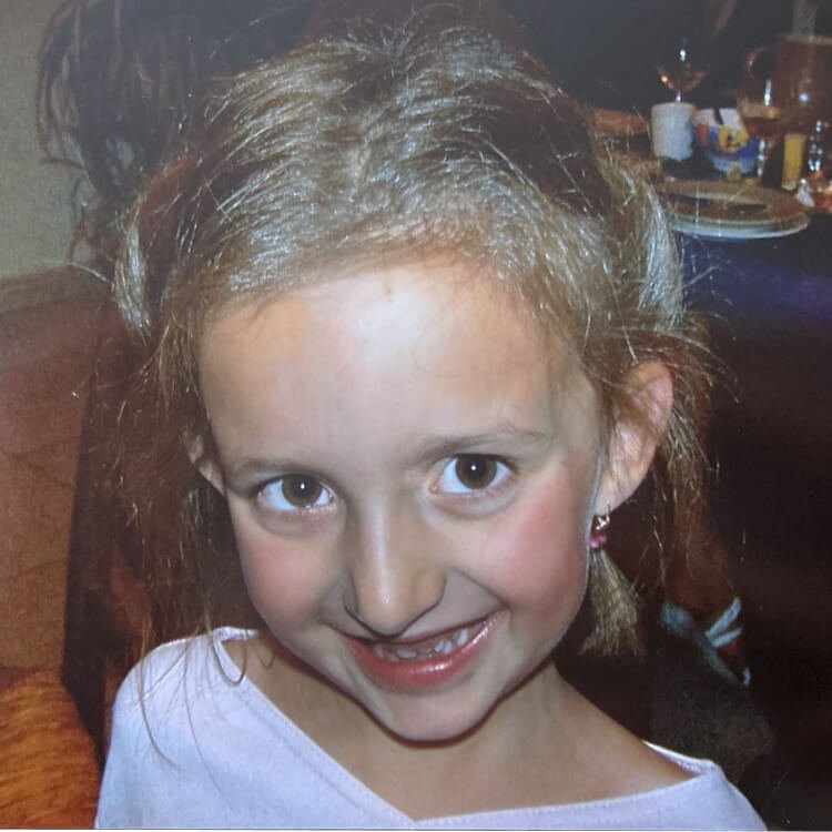 Une jeune fille souriante avec des yeux marron, des cheveux bouclés et une dent manquante.