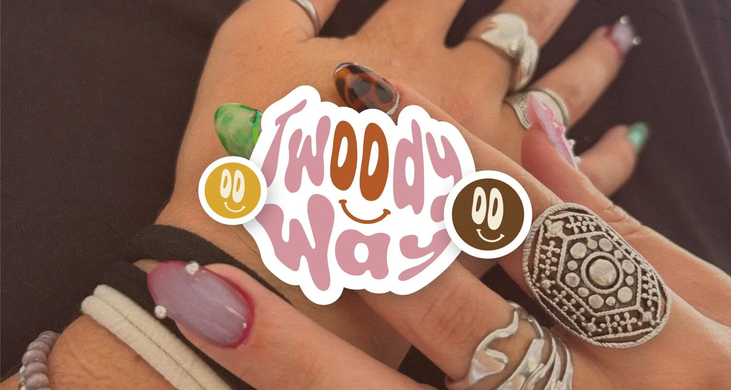 Main avec plusieurs bagues, certains ongles peints en différentes couleurs, et une créature artificielle avec des motifs colorés. Une affiche ou un autocollant avec le texte "Twoody Way" et des smileys souriants couvre une partie de la main.