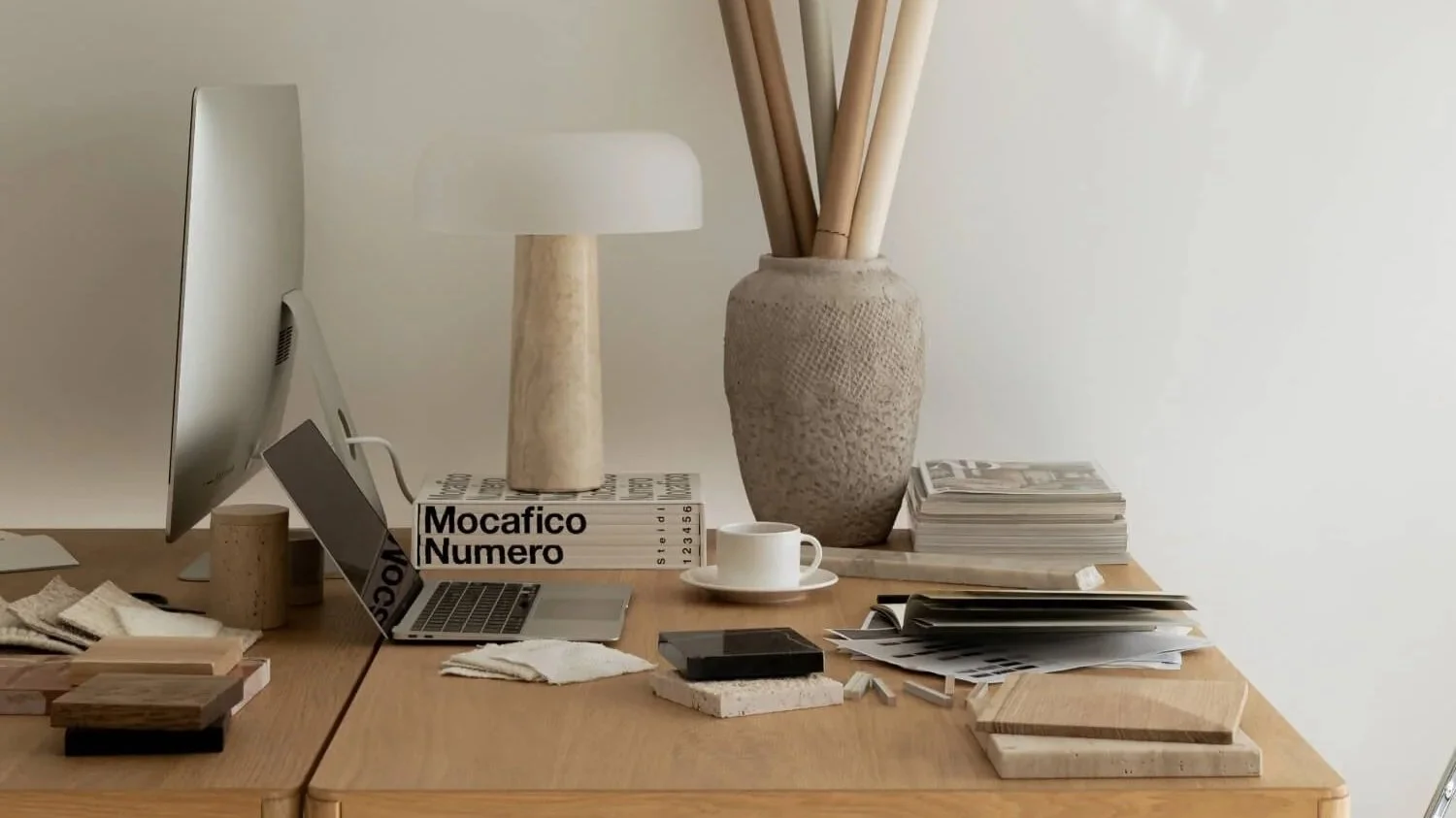 Bureau minimaliste en bois avec ordinateur, lampe blanche, vase en céramique, tasse de café et magazines.