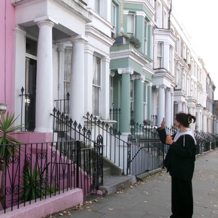 Salomé Dupin, une femme debout sur le trottoir, prenant une photo avec son smartphone devant une rangée de maisons colorées avec des balcons et des colonnes.