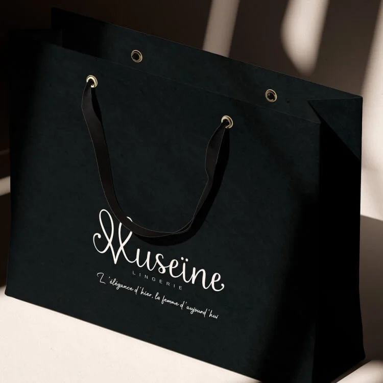 Sac cadeau noir avec poignées en satin, marqué du logo 'Musseine Lingerie' et d'une phrase en français.