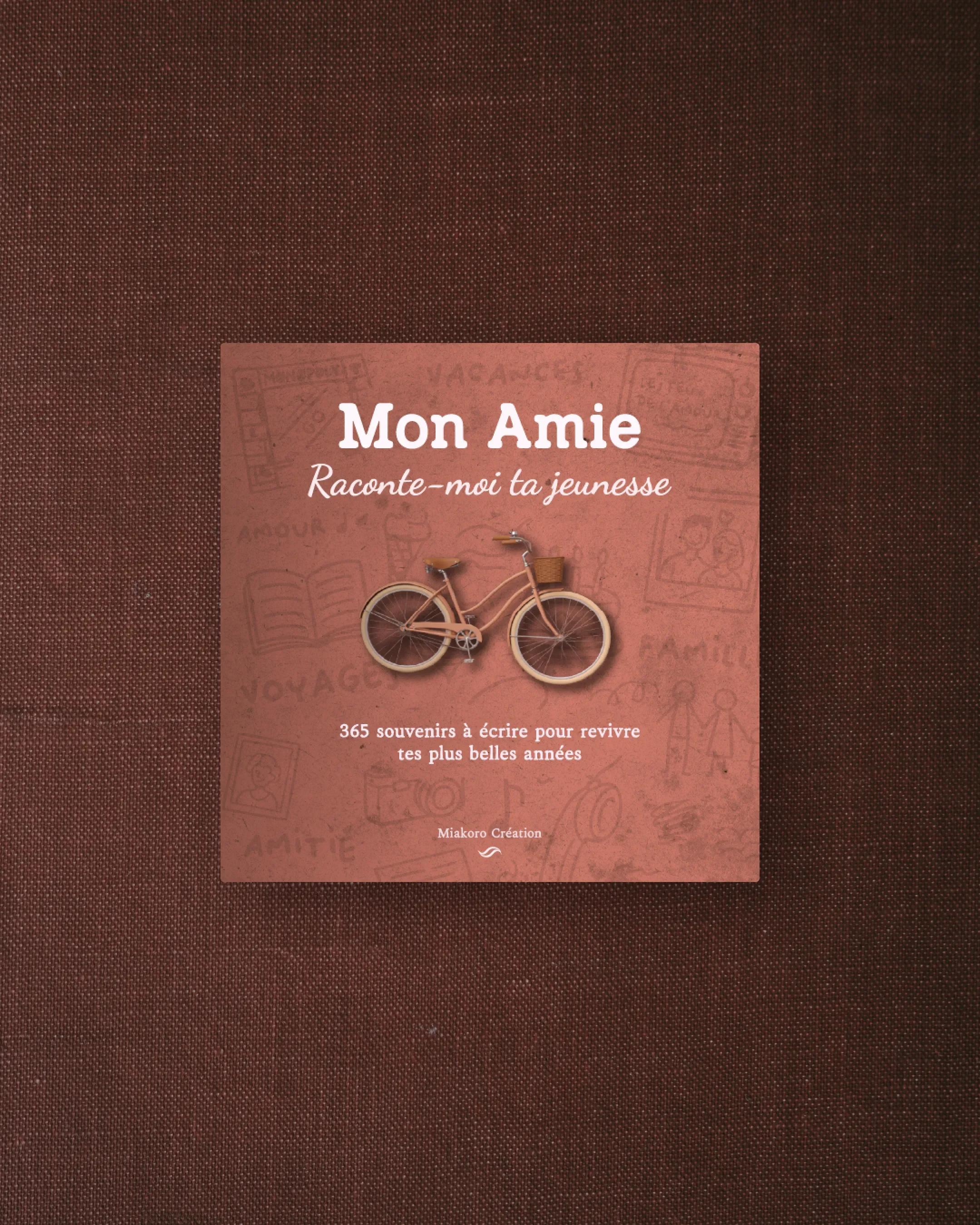 Couverture d'un livre intitulé "Mon Amie" avec un vélo miniature en bois sur la couverture. Le fond de la couverture est rose avec des dessins d'activités et de moments de jeunesse, et le texte indique que le livre contient 365 souvenirs pour revivre