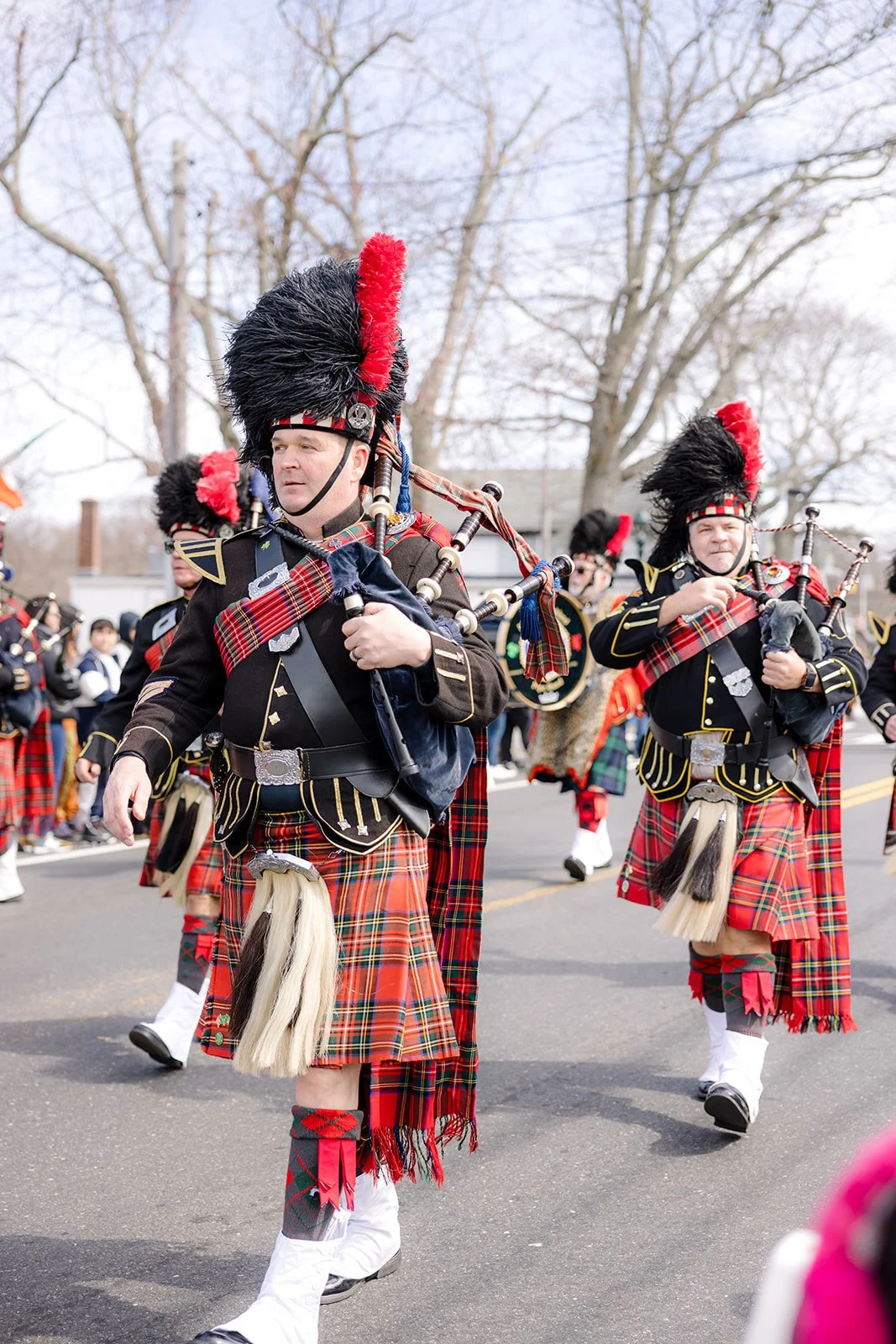 A group of people dressed in traditional Scottish Highland dress, including tartan kilts and feathered hats, participating in a parade and playing bagpipes.