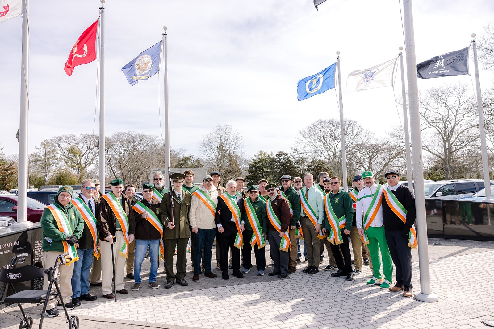 Group of people wearing sashes standing outdoors under flagpoles with flags, in front of parked cars and leafless trees.