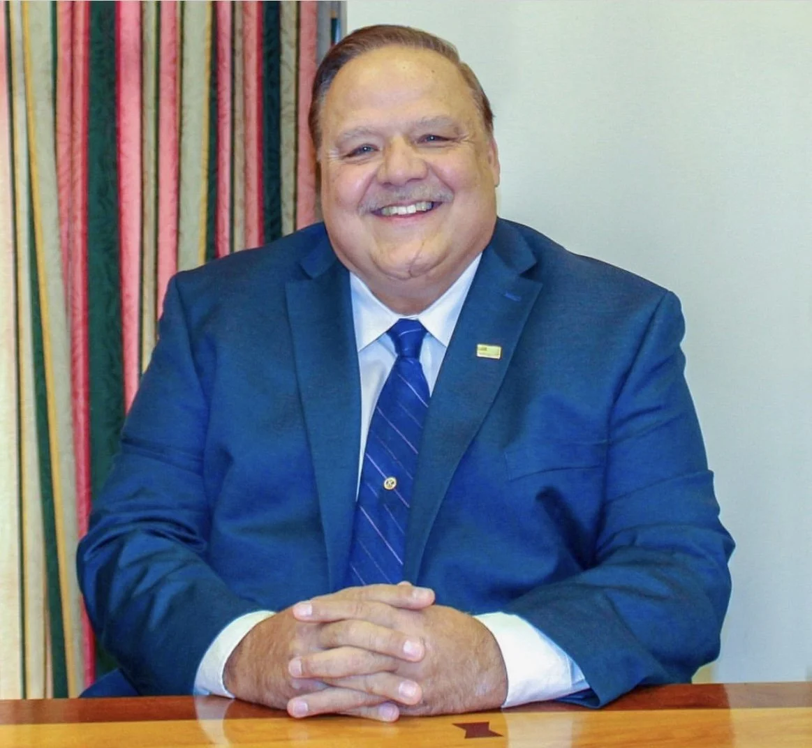 A man in a blue suit and tie, smiling, sitting at a wooden table in front of a colorful striped curtain.