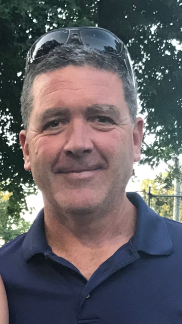 A middle-aged man with short dark hair, wearing sunglasses on his head, smiling slightly, standing outdoors with trees in the background.