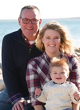 A smiling family of three: a man, a woman, and a young child, outside near water with a clear sky.