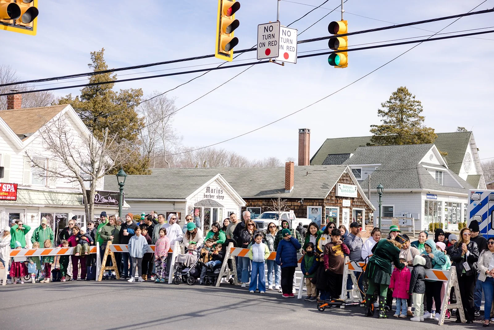 A crowd of people standing behind orange and white barricades on a street, with traffic lights and traffic signs overhead, in a small town with houses and shops in the background.