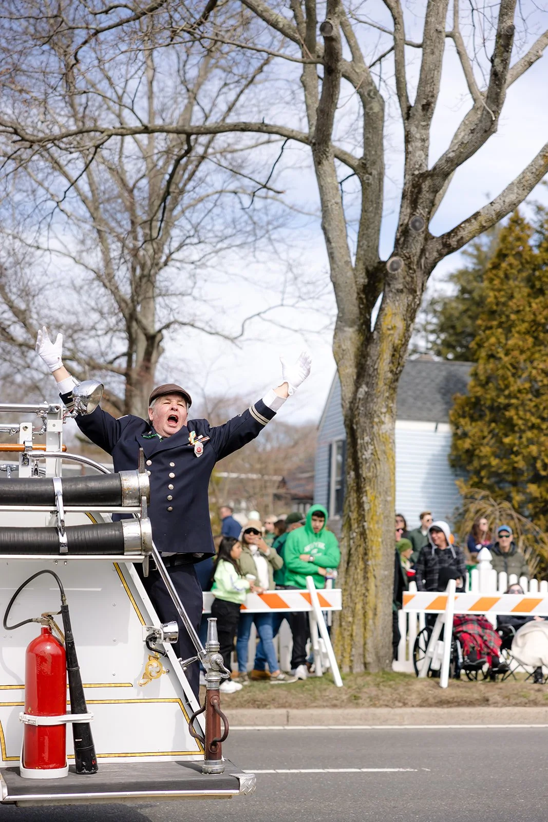 A firefighter in formal uniform on a parade float with arms raised, with spectators in the background watching along a street.