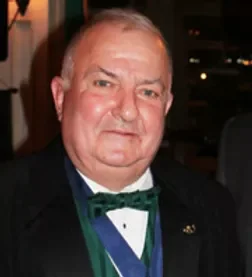 A middle-aged man with short gray hair dressed in formal attire, including a tuxedo with a green bow tie, at an indoor event.