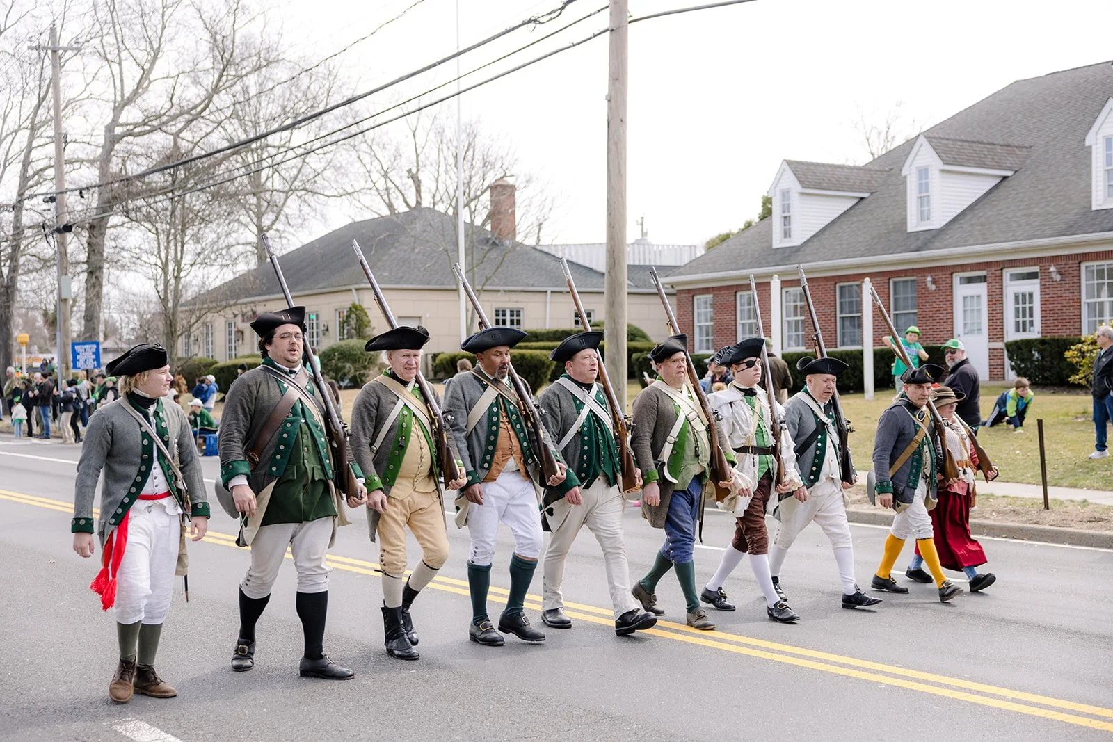 People dressed in historical military costumes participating in a parade, marching on a street with houses and spectators in the background.