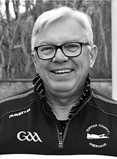 Smiling man with glasses, wearing a sports jacket with GAA emblem, outdoors with trees in background.