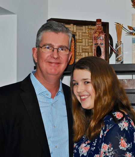A middle-aged man with glasses and gray hair and a young woman with long brown hair standing together indoors, smiling at the camera.