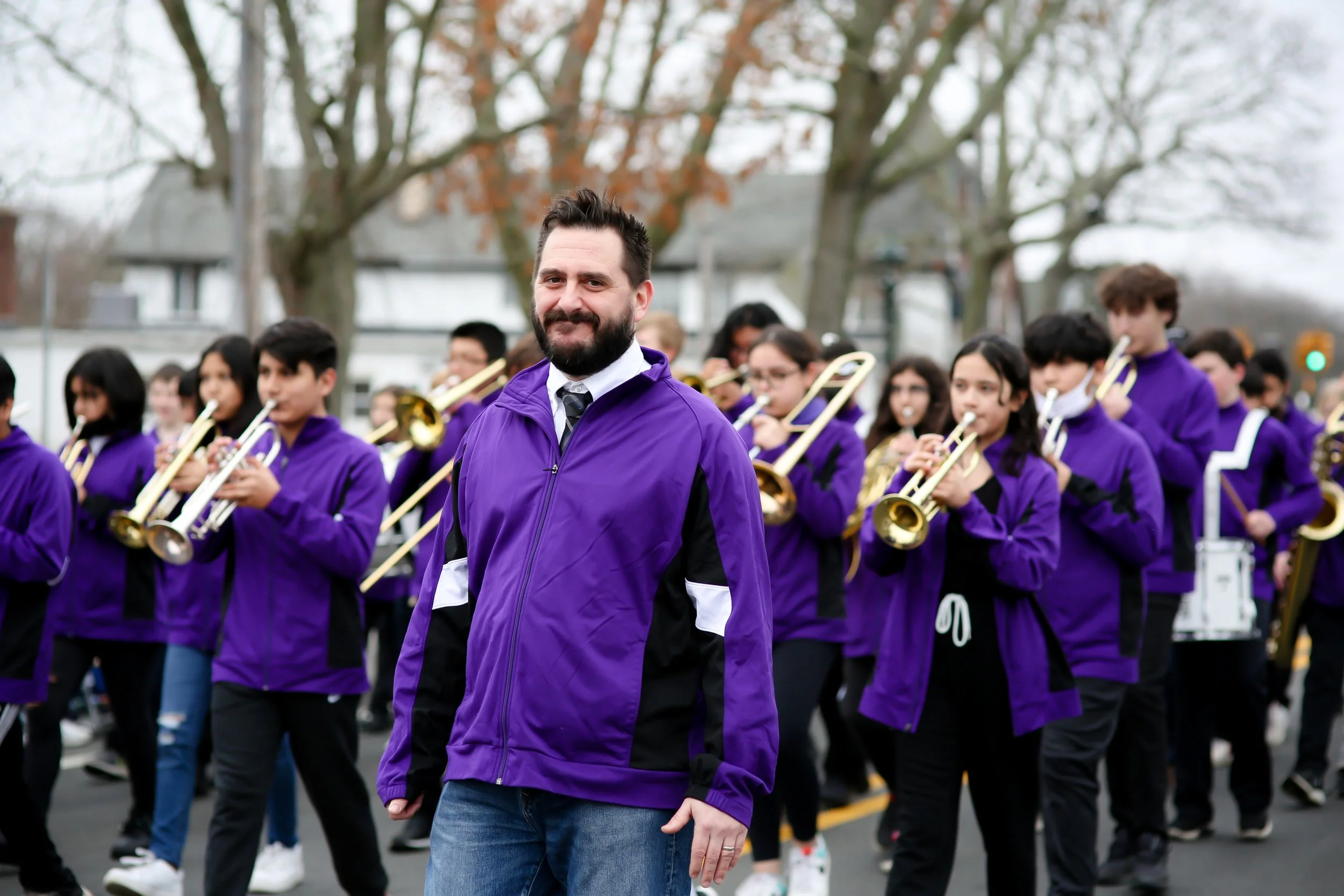 A man with a beard wearing a purple jacket and white shirt standing in front of a marching band with children playing brass instruments during a parade in an outdoor setting with trees and houses in the background.