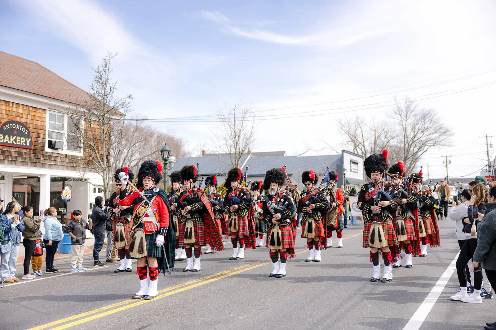 A parade featuring a marching band dressed in traditional Scottish Highland dress with kilts, fuzzy hats, and musical instruments. Spectators line the street watching the performance during daytime.