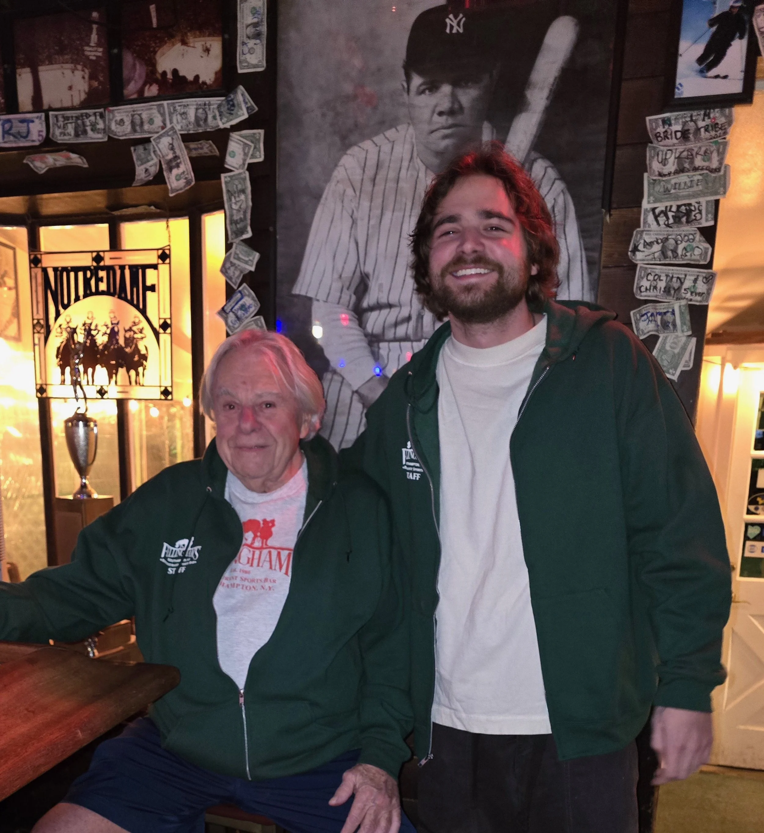 Two men, one elderly and one young, smiling and posing together inside a sports bar. The elderly man has white hair and is wearing a dark green jacket over a gray t-shirt, while the younger man has brown hair and a beard, and is wearing a green jacket over a white t-shirt. Behind them is a large black-and-white poster of a young baseball player in a striped uniform holding a bat, surrounded by dollar bills and memorabilia.