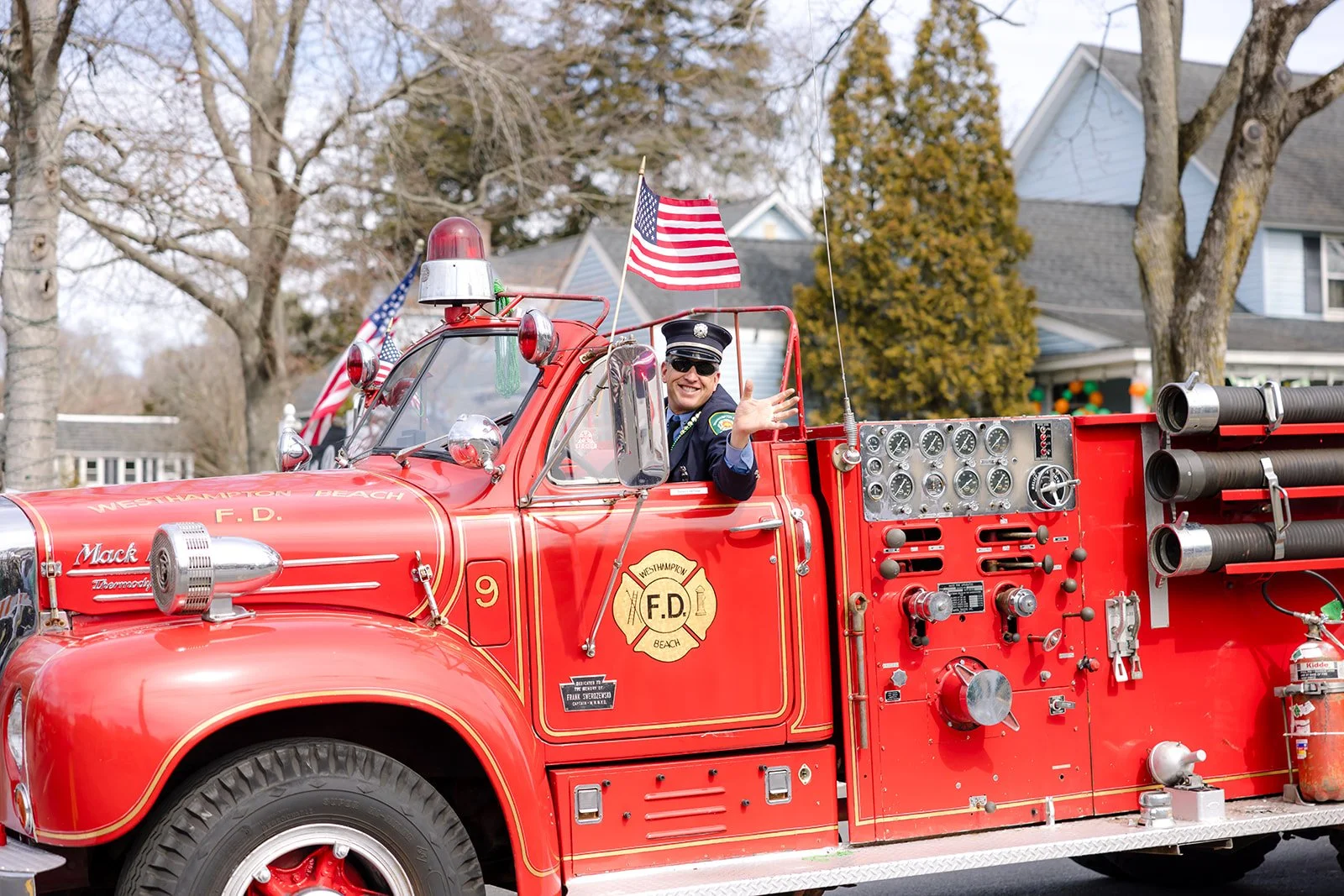 A smiling firefighter in uniform and sunglasses waving from the driver’s seat of a vintage red fire truck decorated with American flags.