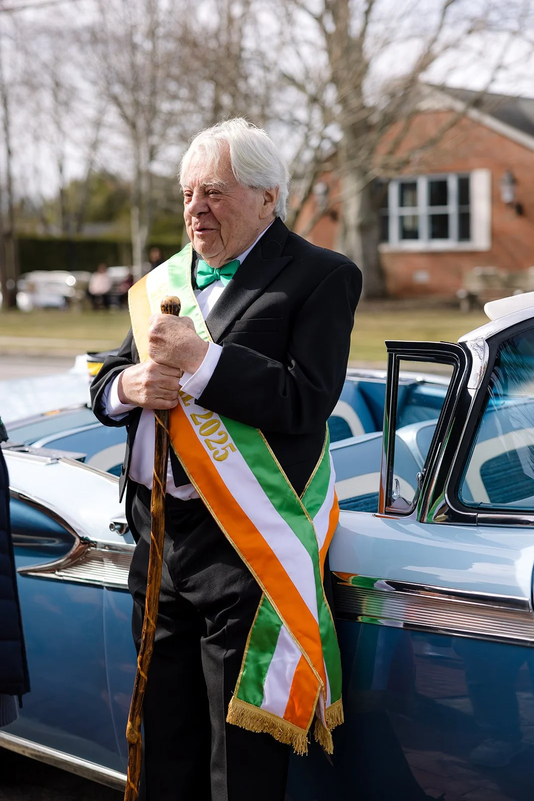 An elderly man dressed in a black tuxedo with a green bow tie stands beside a vintage blue car. He wears a tricolor sash with the year 2025 and holds a wooden cane, smiling as he is at an outdoor event, possibly a celebration or parade.