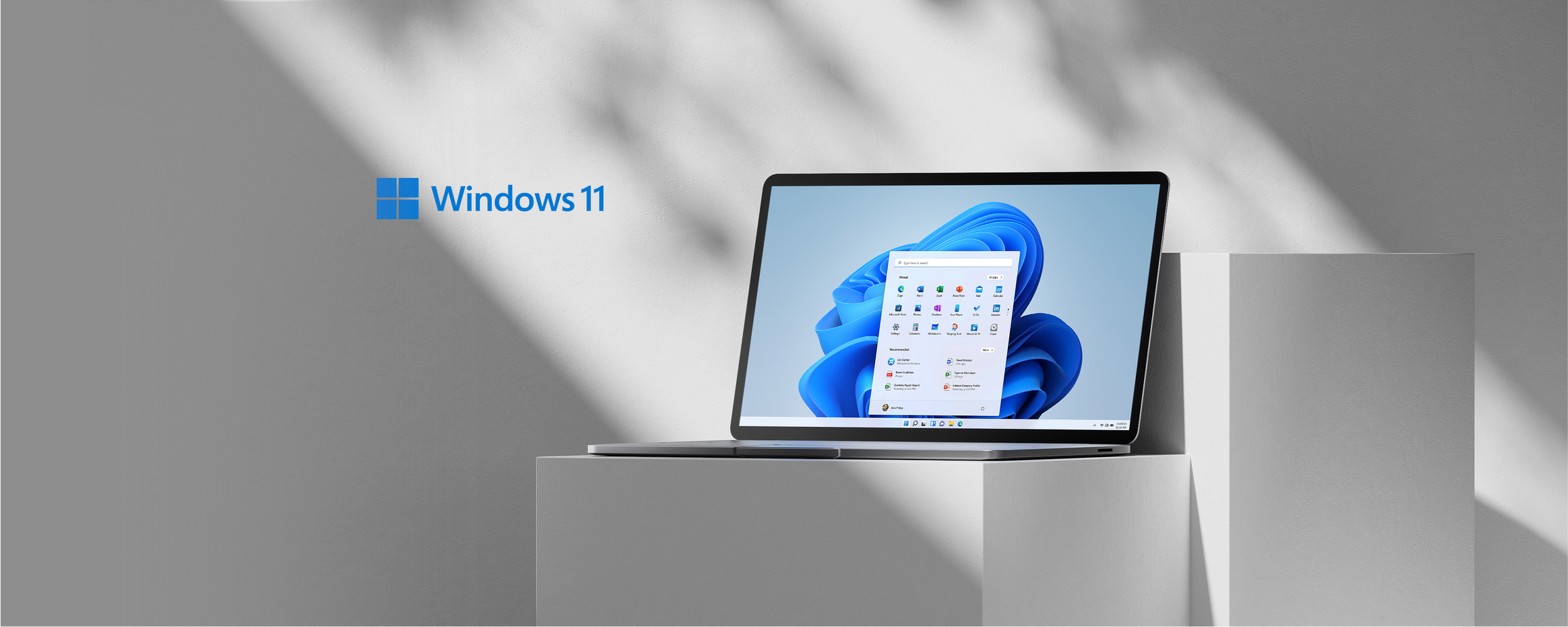 A Microsoft Surface laptop displaying the Windows 11 desktop on a white pedestal against a gray wall with sunlight shadows, with a Windows 11 logo in the upper left corner.