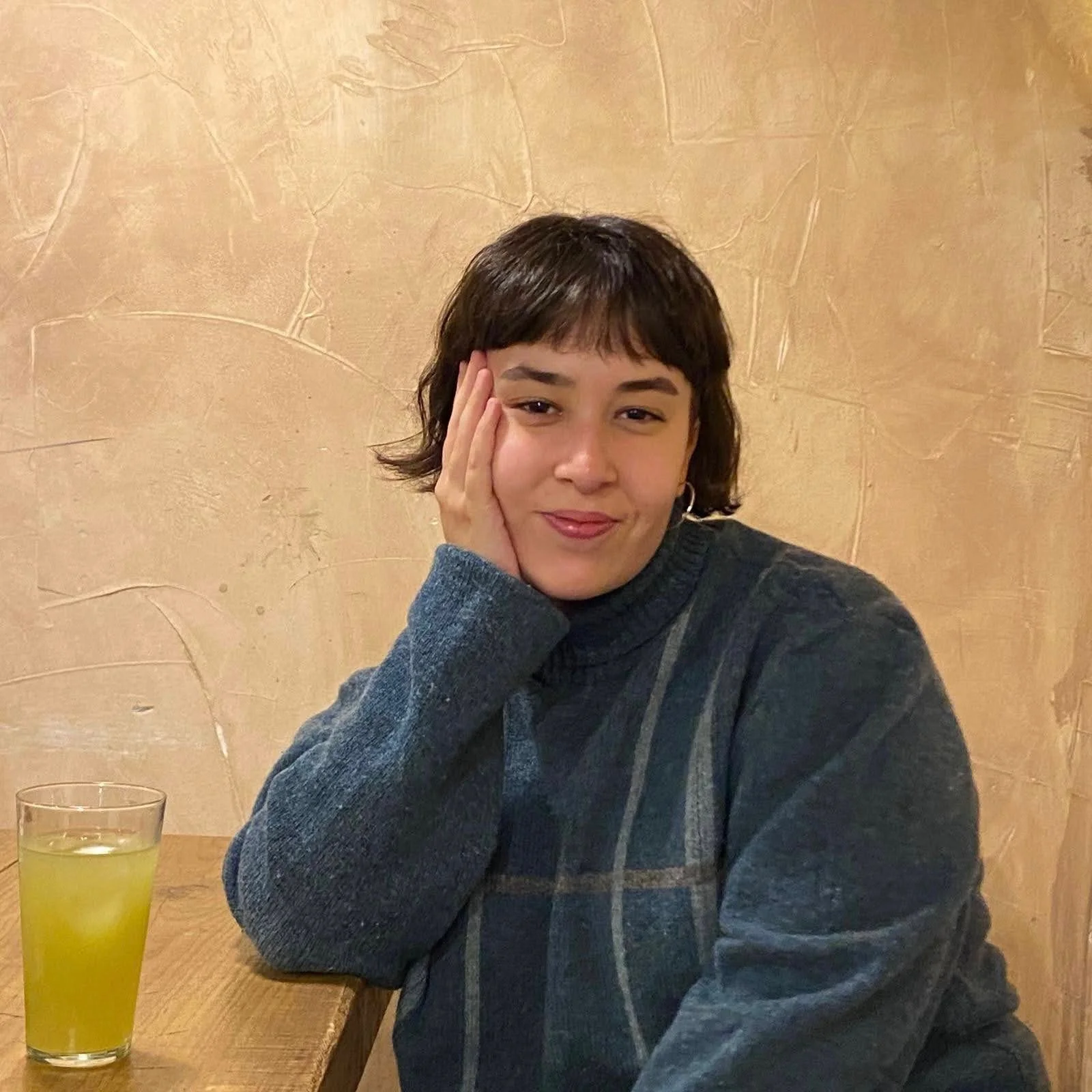 A woman with short dark hair and earrings sitting at a wooden table with a glass of yellow-green beverage, against a textured beige wall background.