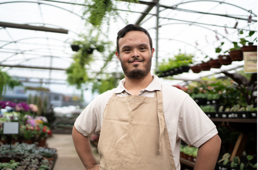 A young man with Down syndrome standing in a greenhouse among flowers and plants, wearing a beige apron over a light-colored shirt, smiling at the camera.