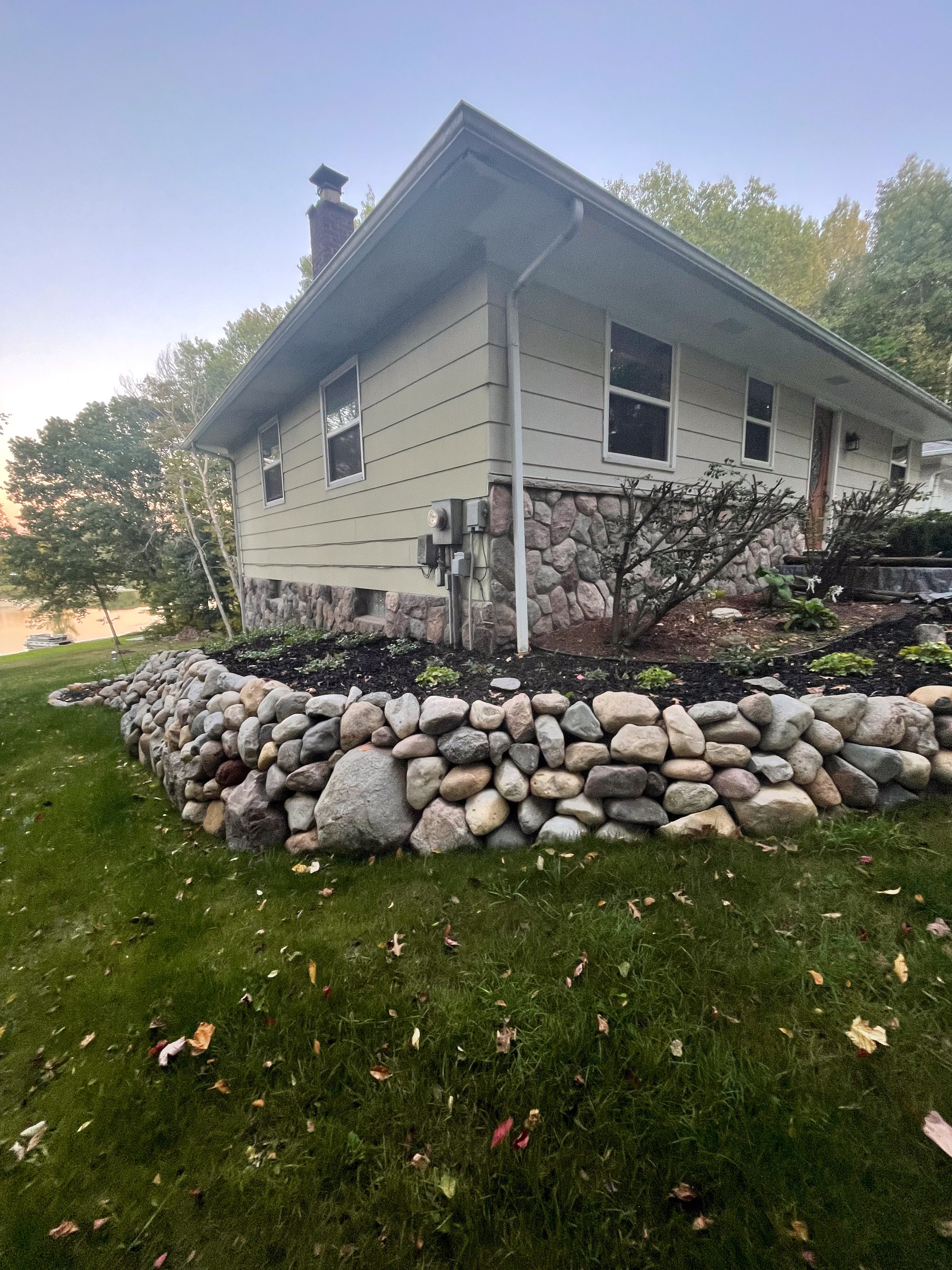 Residential backyard garden with gray stone stairs, a stone-bordered dry creek bed filled with gravel, and a split-level house in the background surrounded by trees.